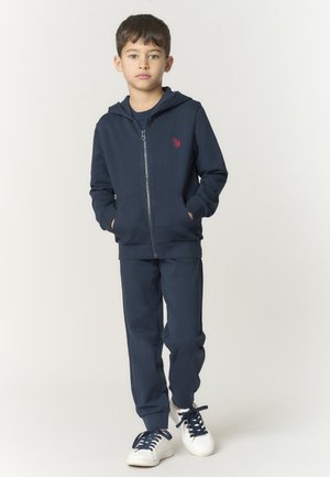 Ragazzo in piedi con le mani in tasca, indossa una felpa con cappuccio blu navy con cerniera, pantaloni coordinati e sneakers bianche con lacci blu navy, su sfondo neutro.