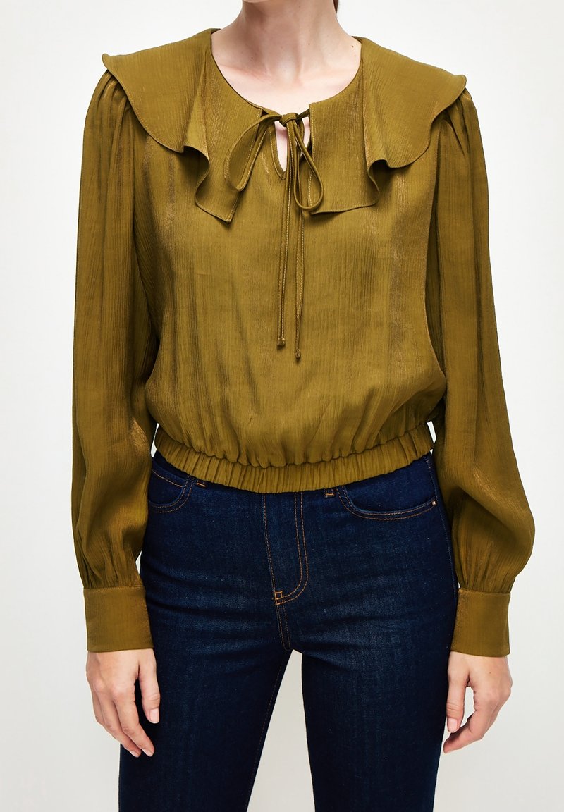 Blouse vert olive avec grand col à volants, manches longues bouffantes, lien au niveau de l'encolure, taille cintrée, portée avec un jean bleu foncé taille haute.