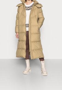 Abrigo largo acolchado beige con cierre de cremallera, cuello alto y textura acolchada. Llevado sobre un suéter crema y pantalones marrones, combinado con botas blancas.