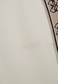 Tissu de couleur crème avec le logo "GUESS" en relief. Le design comprend des accents contrastants et une surface texturée.