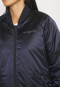 Mørkeblå bomberjakke lavet af skinnende materiale med lynlås foran, ribbet krave og hvidt logo-tekst "TOMMY HILFIGER EST. 1985" på brystet.