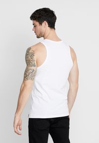 Hvit bomulls tank topp med rund hals, ermeløs design og rett nederkant. Har en tatovering på venstre arm, svarte jeans.