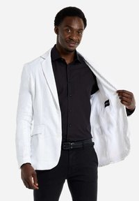 Weißer Pailletten-Blazer mit strukturiertem Oberflächen und zwei Fronttaschen, getragen über einem schwarzen Hemd und einem schwarzen Gürtelhose. Futter ist weiß.