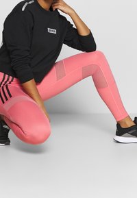 Sweatshirt noir avec des accents argentés et un logo, associé à des leggings roses clairs avec des panneaux en maille et des détails en bandes noires, portés avec des baskets.