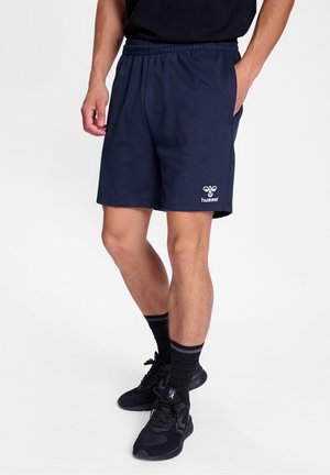 Männliches Model trägt marineblaue Hummel-Shorts mit elastischem Bund, schwarze Sportschuhe, schwarze Socken, steht vor weißem Hintergrund.