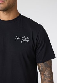 Czarny bawełniany t-shirt z krótkimi rękawami z białym, kursywnym napisem "Closure London Studios" na lewej piersi.