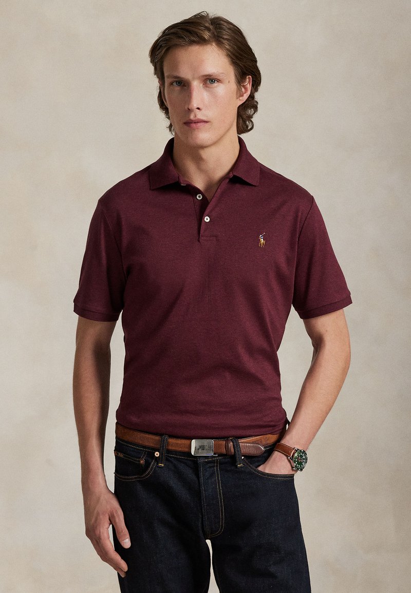 Ung mand med mellem langt hår iført en bordeaux polo-shirt, mørke jeans, brun bælte og et ur med grøn urskive.