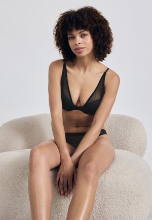 Musta verkosta valmistettu bralette ja alushousut, joissa on ohut olkaimien muotoilu, V-kaula-aukko ja sileä pinta. Ne ovat sijoitettu vaaleanväriselle, teksturoidulle tuolille.