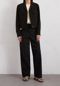 Svart kort jacka med krage och fyra framfickor, kombinerad med högmidjade svarta byxor. Bärs med svarta platta sandaler.