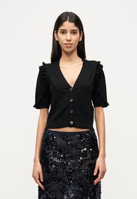 Μαύρο κοντό ζακέτα με κοντά μανίκια, διακοσμητικά βολάν και πέντε κουμπιά. Συνδυασμένο με μια λαμπερή μαύρη φο ύστα με παγιέτες.