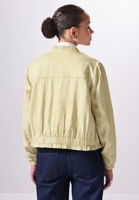 Giacca gialla chiara con vita arricciata, polsini elastici e una texture liscia. Presenta un semplice colletto e un design con spalle a yoke sul retro.