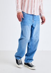 Homme portant un jean bleu clair ample, des baskets noires et blanches, et une chemise à manches longues rayée rose et blanche debout sur un sol blanc.