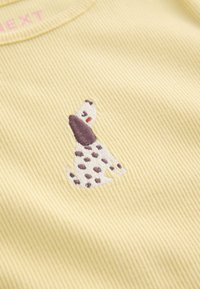 Žuti tkanina s rebrastim uzorkom s izvezenim motivom smeđeg i bijelog psa s mrljama. Ima okrugli izrez i mekanu teksturu.