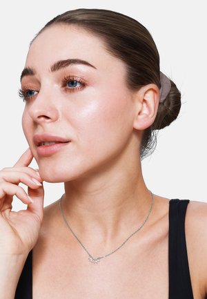 Zilveren ketting met een kleine bloemenhanger, gedragen op blote huid, tegen een zwarte top. Haar is in een knot gestyled, met natuurlijke make-up.
