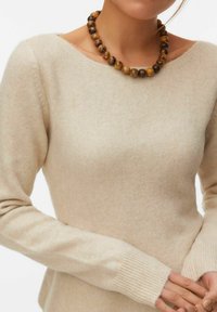 Pull beige à manches longues avec poignets côtelés, porté avec un collier chunky de perles rondes en bois sur une personne les mains jointes.