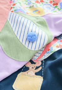Kolorowy patchwork z tkaniny z miękkimi strukturami, niebieskim filcowym pomponem, wzorem w paski oraz haftowaną myszką w detalu kieszonki.