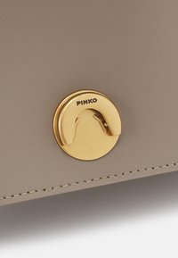 Dettaglio di una borsa in pelle tan con un hardware circolare color oro con la parola "PINKO" incisa, che mette in risalto una texture liscia e cuciture precise.