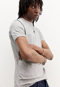 Tricou gri cu mânecă scurtă, în croi slim, cu guler rotund și textură moale. Accesorii remarcabile includ brățări din argint și un colier cu pandantiv.