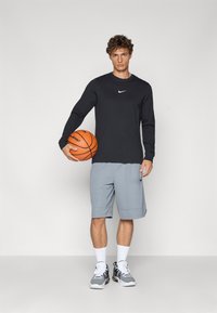 Sort langærmet Nike skjorte og grå shorts. Holder en basketball. Hvide sneakers med sorte detaljer og sokker. Enkel, sporty design.