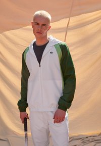 Balta un zaļa kapucīna jaka ar rāvējslēdzēju, ar garām piedurknēm un Lacoste logo. Uzvilkta virs tumšas krekli, uz pelēka fona.
