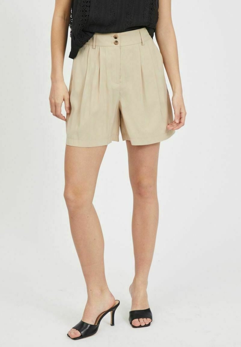 Femme portant un short plissé beige taille haute avec deux boutons, un haut noir sans manches et des mules noires à talons ouvertes à l’avant.