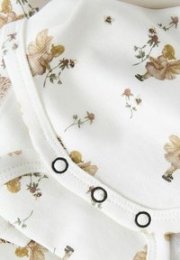 Vit tyg med blommönster och prinsesstryck, med svarta knappar och texturerad ribbing längs kanterna. Mjukt, lätt material.