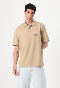 Μπεζ πόλο μπλούζα από βαμβάκι με γιακά, κοντά μανίκια και τετράγωνο λογότυπο στο αριστερό στήθος. Φορεμένη με ανοιχτό μπλε τζιν.