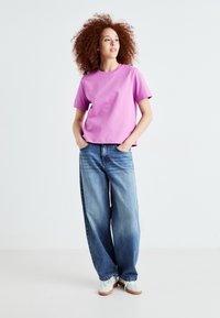 T-shirt in cotone rosa con maniche corte, abbinato a jeans a gamba larga blu chiaro. Il modello indossa sneakers bianche, in piedi con le mani nelle tasche.