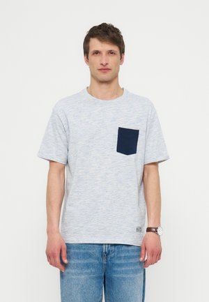 Άνδρας στέκεται προς τα εμπρός φορώντας ανοιχτογκρι κοντομάνικη μπλούζα με σκούρο μπλε τσέπη στο στήθος και μπλε τζιν, με απλό λευκό φόντο.