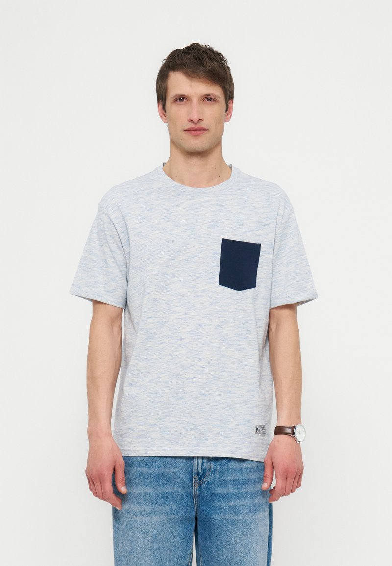 Άνδρας στέκεται προς τα εμπρός φορώντας ανοιχτογκρι κοντομάνικη μπλούζα με σκούρο μπλε τσέπη στο στήθος και μπλε τζιν, με απλό λευκό φόντο.