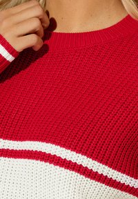 Punainen neulepaita, jossa on teksturoitu kuvio, valkoiset vaakaraitat kauluksessa ja hihoissa. Lähikuva kankaan kudontasta.