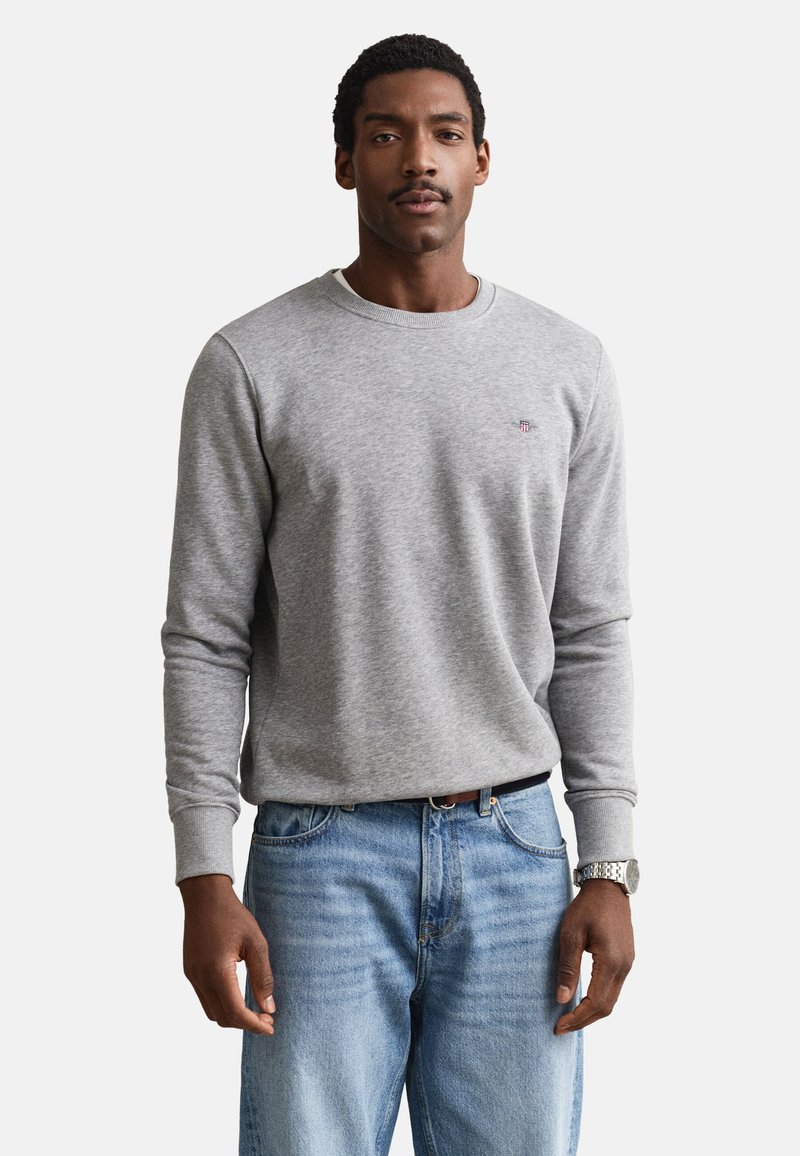 Homme portant un pull gris clair à manches longues et col rond, ainsi qu'un jean bleu, debout les mains détendues le long du corps.
