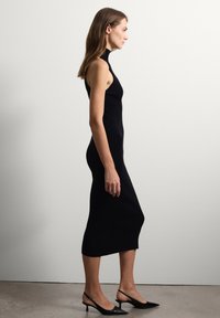 Robe midi noire en tricot côtelé avec un col haut, une silhouette ajustée et une fente sur le côté. Portée avec des talons noirs pointus.