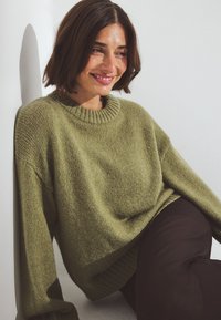 Oliwkowy sweter robiony na drutach z okrągłym dekoltem, opuszczonymi ramionami i ściągaczem u dołu. Stylizacja z ciemnymi spodniami, na tle białej ściany.