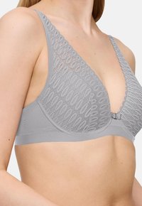 Ženska v svetlo sivi čipkasti nedrček s sprednjim zapiranjem, ki prikazuje zgornji del trupa in podroben čipkast vzorec na skodelicah in naramnicah.