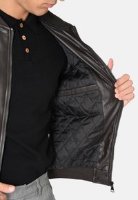 Giacca in pelle marrone con un interno trapuntato nero. Presenta una zip completa, polsini a coste e una finitura liscia. Esibita aperta.