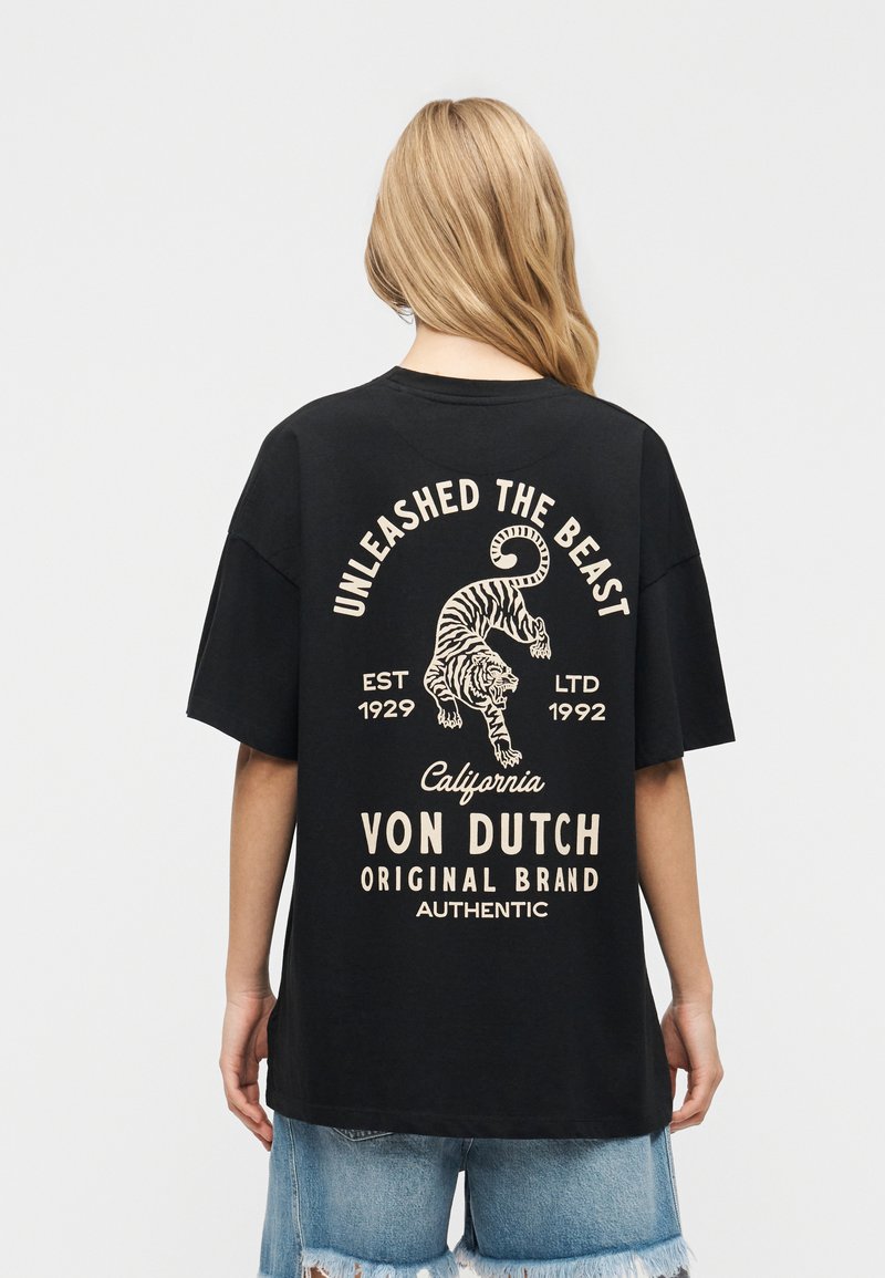 Άτομο με μακριά ξανθά μαλλιά φοράει μαύρο T-shirt με μπεζ γραφικό τίγρης και κείμενο "Απελευθέρωσε το Θηρίο" και "Von Dutch Original Brand."