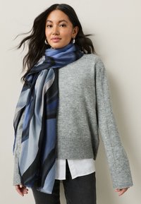 Gray knitted sweater with ribbed cuffs, layered over a white shirt. A blue and black abstract patterned scarf drapes around the neck.