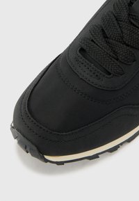 Black sneaker with a smooth, textured upper, featuring a raised toe cap and thick black laces, and an off-white midsole accent.