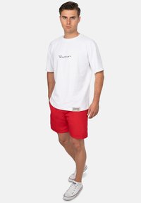 T-shirt bianca in cotone con logo nero sul petto, abbinata a pantaloni corti rossi. Tessuto leggero con una texture liscia. Sneakers bianche.