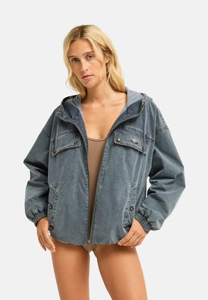 Blonde vrouw die een losse, vervaagde blauwe jas met rits en klepzakken draagt over een beige top, staand tegen een eenvoudige lichte achtergrond.
