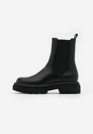 Botte Chelsea en cuir noir avec panneaux latéraux élastiques, semelle crantée épaisse, et languette à l'arrière, présentée de profil sur fond blanc.