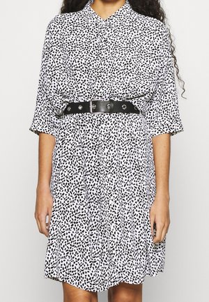 Kobieta nosząca białą sukienkę koszulową w czarne nieregularne kropki, z talią podkreśloną szerokim czarnym paskiem z srebrną klamrą.