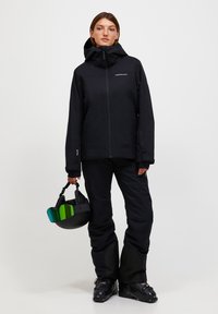 Giacca da sci nera con cappuccio, abbinata a pantaloni coordinati. Il modello tiene in mano un casco nero con visiera verde e blu. Design elegante e funzionale.
