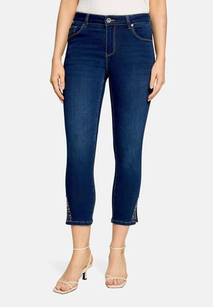 Slim fit mørkeblå korte jeans med forreste knap, lommer og rhinsten detaljer ved ankelåbninger, båret med beige hælede sandaler.