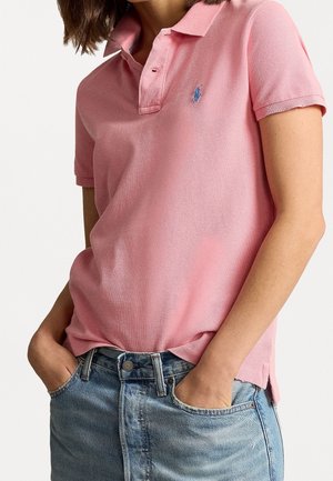 Vrouw die een roze polo met korte mouwen en lichtblauwe spijkerbroek draagt, handen in de zakken, bijgesneden ter hoogte van nek en heupen.