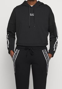 Černá mikina s kapucí a odpovídající džínsy vyrobené z měkké tkaniny. Obsahuje bílé logo a pruhy podél rukávů a nohavic, s nastavitelnou šňůrkou.