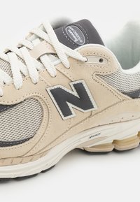 Beige sportskor med grått nät och mockadetaljer. Har vita snören, en framträdande "N"-logotyp och en vadderad sula. Texturerade designelement.