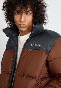 Chaqueta acolchado con la mitad inferior marrón y la mitad superior negra. Hecha de un material brillante y acolchado. Presenta un alto collar y cierre de cremallera.