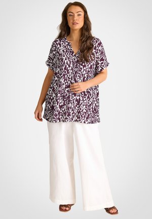 Frau trägt eine locker sitzende Bluse mit kurzem Arm in lila-weißem Muster, weiße Hosen mit weitem Bein und braune Sandalen mit offenem Zehenbereich, steht vor einfachem Hintergrund.